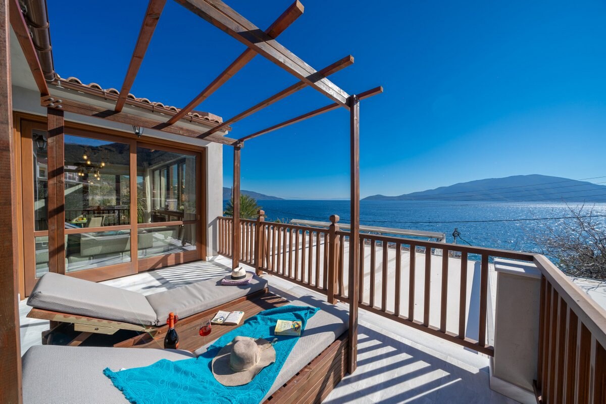 A sunny balcony offers lounging space with two comfortable chaise lounges and a textured blue blanket. A table with a bottle of wine and snack plates sits nearby, while sweeping sea views and distant hills create a serene backdrop under a clear blue sky.