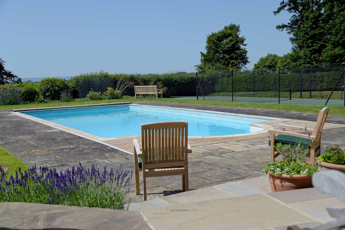 An outdoor swimming pool is showcased surrounded by a paved patio. Two wooden chairs are positioned nearby, offering a space to relax. A well-maintained lawn and hedges are visible, with a tennis court in the background. Blue skies create a bright and inviting atmosphere.