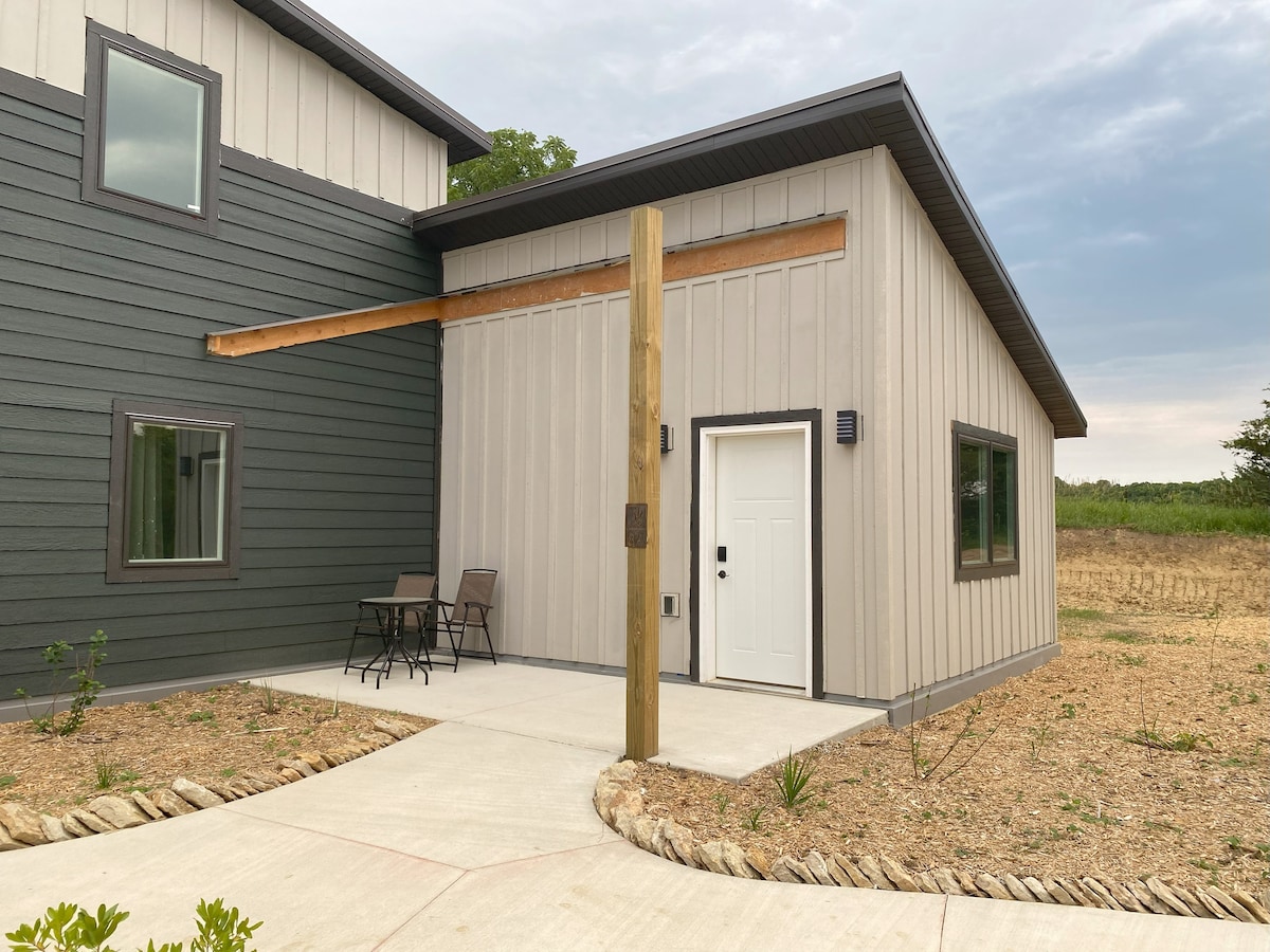 An exterior view of the rental unit presents a modern design with a mix of dark gray and light beige siding. A covered entryway features a wooden beam and leads to a white door. Two outdoor chairs and a small table are positioned on a paved walkway surrounded by gravel landscaping.