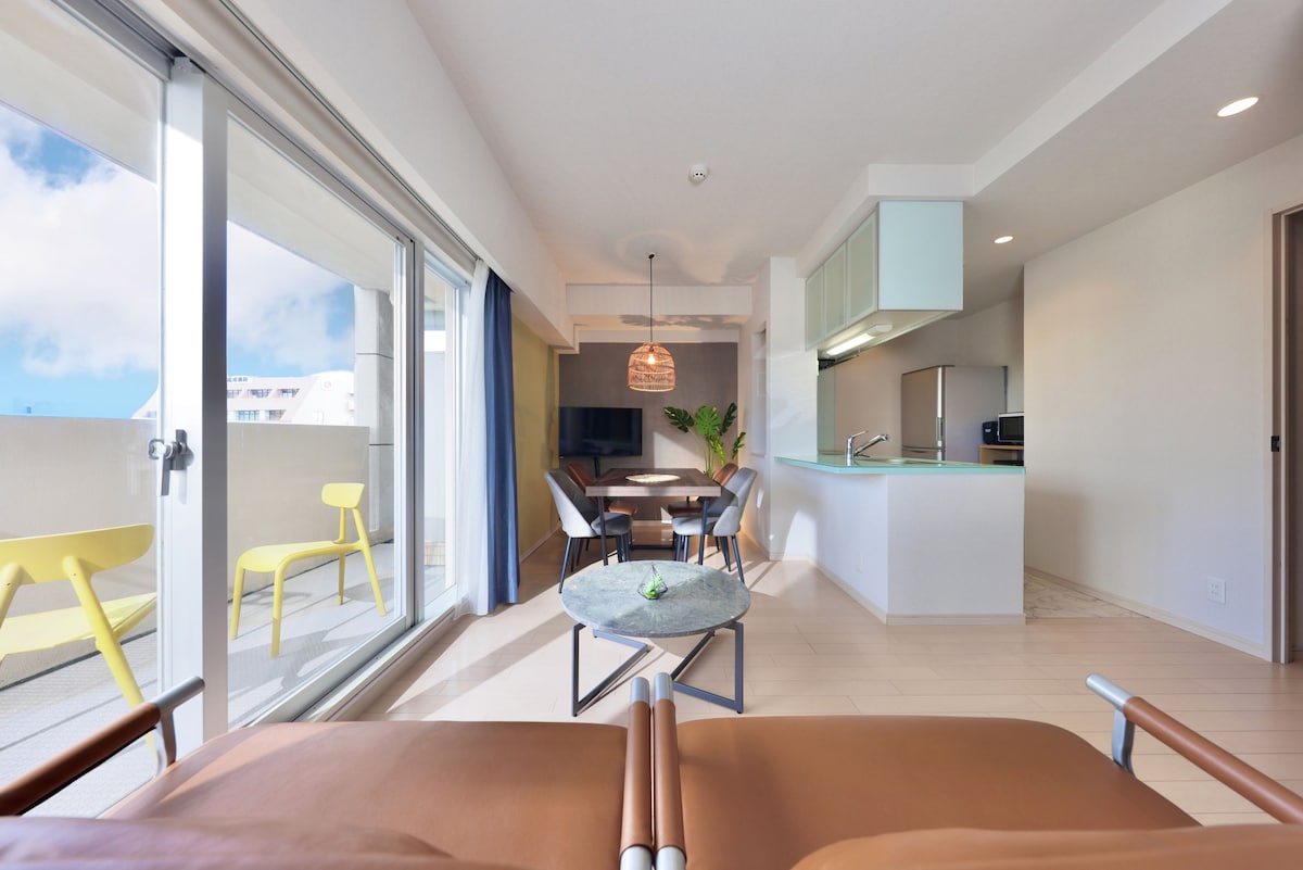 The open living area features large windows that fill the space with natural light. A comfortable sofa faces a round dining table, with chairs positioned around it. A modern kitchen is visible in the background, showcasing sleek cabinetry and appliances.
