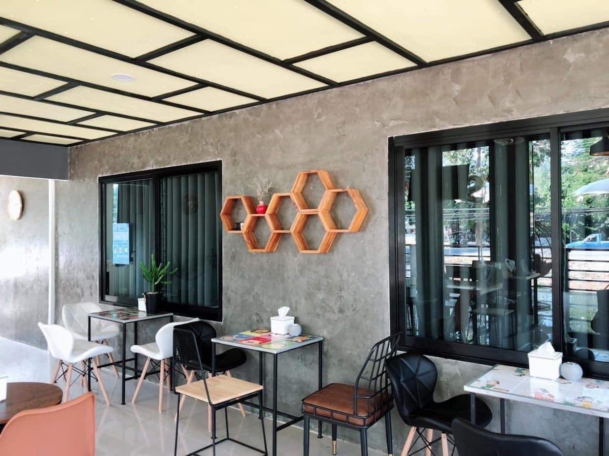 A modern dining area features sleek furniture including a mix of white and black chairs around small tables. Natural light enters through large windows, and a decorative wall shelf in a honeycomb design is accented with plants, creating a refreshing atmosphere.