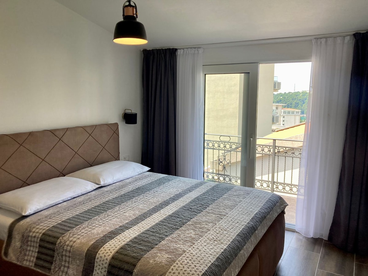 A bedroom is presented with a spacious bed adorned with a patterned blanket. Natural light enters through a glass door that leads to a balcony, framed by sheer white curtains and solid dark curtains. A modern light fixture hangs above the bed.