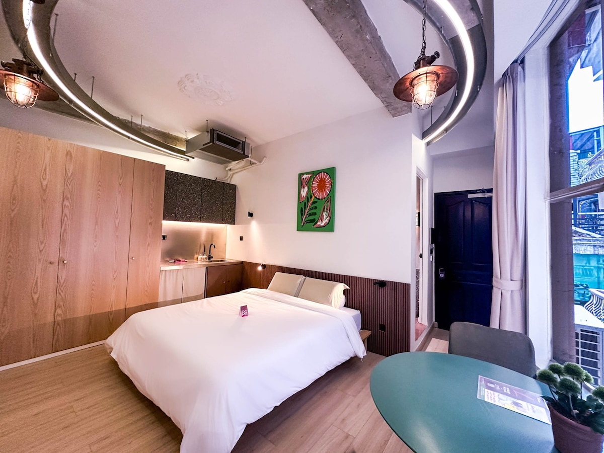 A serene room is presented with a spacious bed centered against wooden cabinetry, providing a minimalist ambiance. Soft light is introduced by stylish fixtures, complemented by modern decor and a vibrant artwork on the wall. A small dining table is positioned beside a large window.