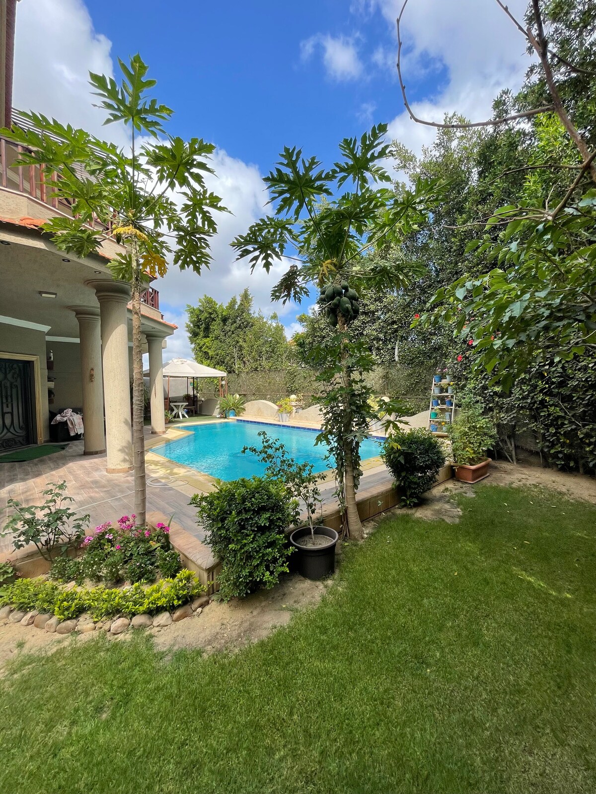 A well-maintained garden surrounds a bright blue swimming pool, with vibrant greenery and colorful flowers enhancing the natural setting. A terrace is visible beside the pool, accompanied by a sunshade for outdoor relaxation, under a partly cloudy sky.