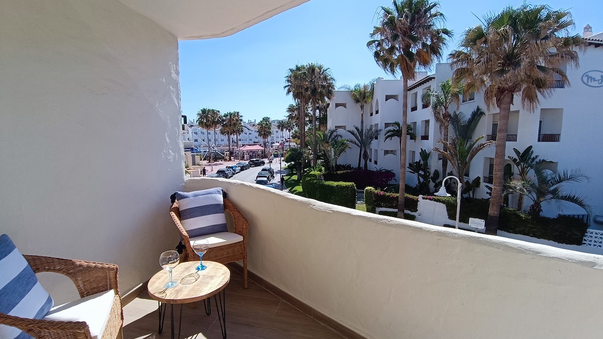 A terrace area features two wicker chairs with striped cushions and a small table, overlooking palm trees and landscaped gardens. Sunlight illuminates the space, creating a relaxed setting. The view includes a pathway and nearby buildings, contributing to the vibrant atmosphere of the surrounding area.