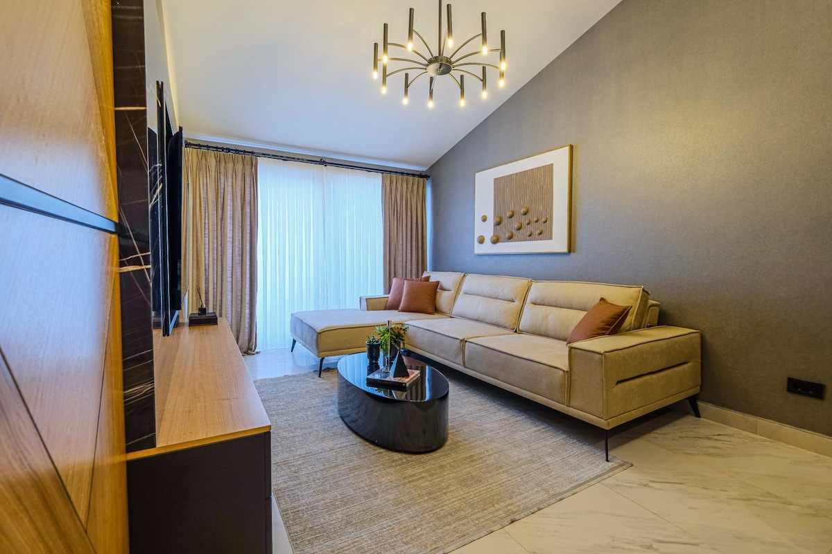 A modern living area is featured with a spacious light beige sofa, complemented by decorative cushions. A sleek black coffee table is centered on a soft rug, while natural light flows in through large windows with sheer curtains. Wall-mounted television adds a contemporary touch.