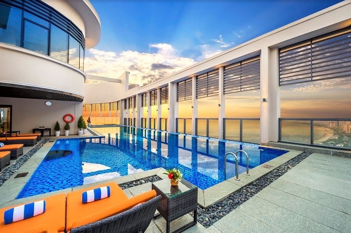 The rooftop swimming pool, located on the 33rd floor, offers expansive views of the sea and surrounding landscape. Lounge chairs in vibrant orange surround the pool, while large windows allow natural light to enhance the space. The tranquil atmosphere is complemented by a colorful sunset sky.