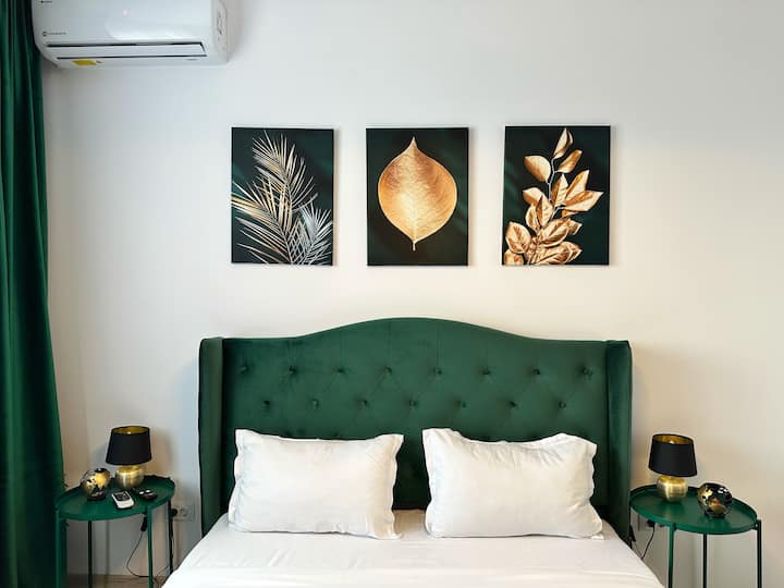 ห้องนอนพร้อมหัวเตียงบุผ้าสีเขียว เครื่องนอนสีขาว โต๊ะข้างเตียงสีเขียวที่เข้ากันกับโคมไฟสีดำ และงานศิลปะพฤกษศาสตร์สีทอง 3 ชิ้นเหนือเตียง