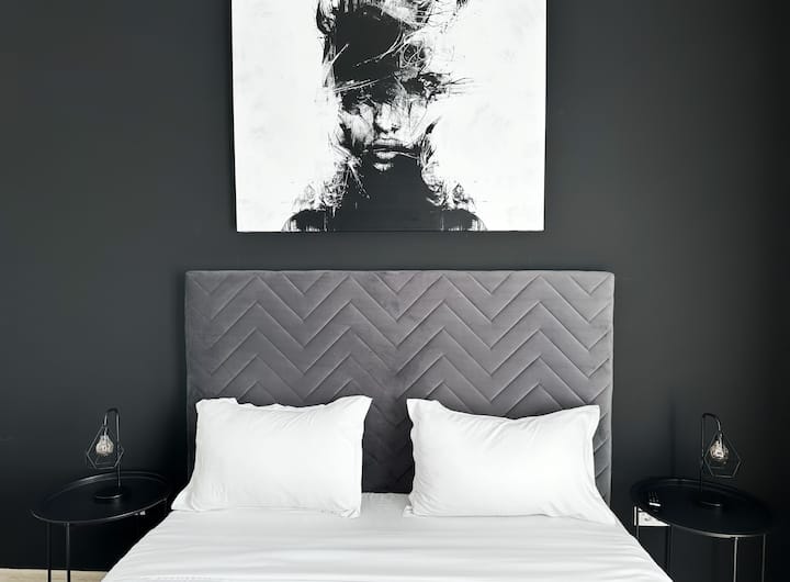 ห้องนอนสไตล์โมเดิร์นพร้อมหัวเตียงบุนวมสีเทา เครื่องนอนสีขาว ผนังสีดำ งานศิลปะสีขาวดำเหนือเตียง และโต๊ะข้างเตียงสีดำพร้อมโคมไฟ
