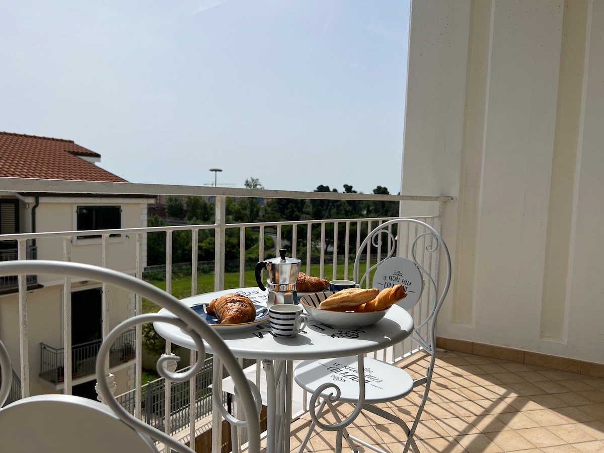 A small balcony features a round table set for breakfast, accompanied by a coffee pot, cups, and fresh pastries. Two white chairs are positioned around the table, while the view includes greenery and nearby buildings, enhancing the relaxing atmosphere.