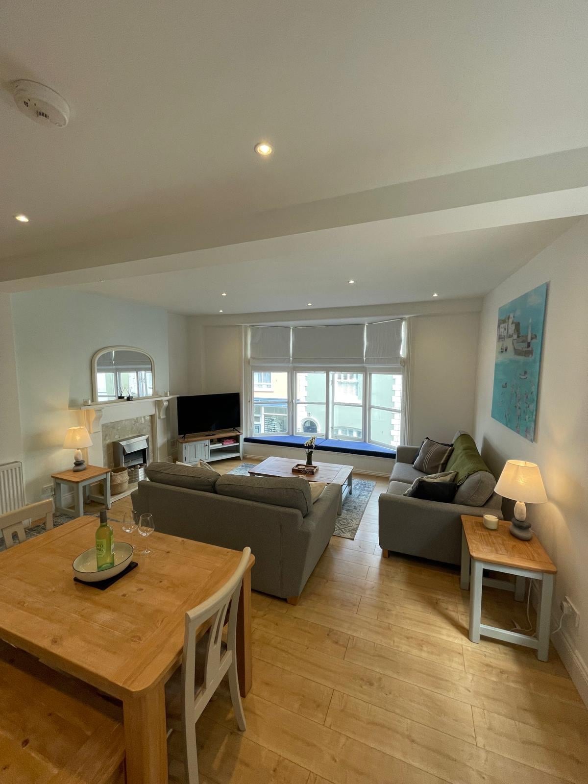 An open-plan living and dining area is visible, featuring a comfortable gray sofa and an inviting armchair positioned around a coffee table. A wooden dining table with two chairs is located nearby. Natural light is streamed through large windows, creating a bright atmosphere.