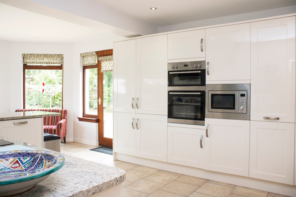A modern kitchen features sleek white cabinetry, including an integrated microwave and multiple ovens. A textured counter is complemented by a decorative bowl. Large windows allow natural light to illuminate the space, providing views of the outdoors. A cozy seating area is seen in the background.