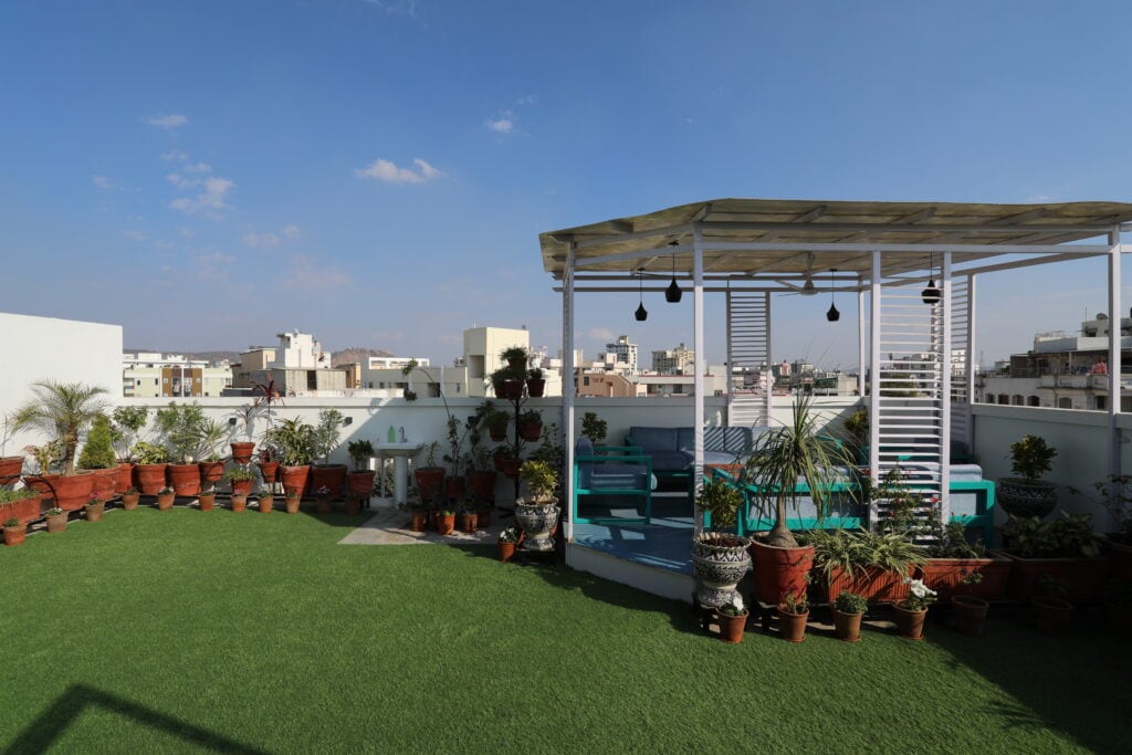A rooftop garden is presented with lush green artificial grass and an array of potted plants. A shaded seating area is visible, accompanied by decorative pots. The surrounding skyline displays an urban landscape, enhancing the outdoor ambiance.