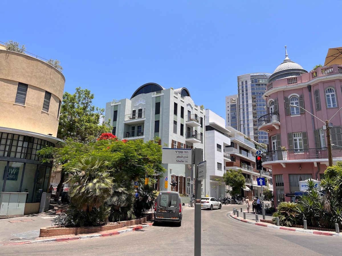 A vibrant street scene showcases a mix of architectural styles, including a modern building with rounded features and a historic structure with a domed roof. Lush greenery surrounds the area, with bicycles parked alongside the road. Signs and traffic signals are visible, indicating the busy atmosphere.