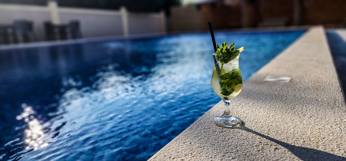 A refreshing cocktail sits on the edge of a large swimming pool, with clear blue water shimmering in the sunlight. The pool's edge is outlined by a textured stone surface, highlighting the drink's vibrant colors.