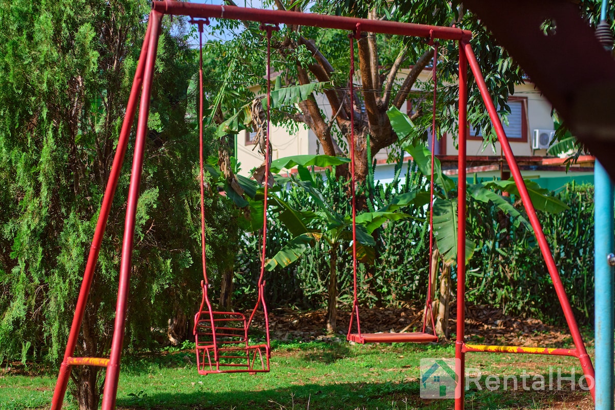 A vibrant red swing set is situated in a lush green yard, surrounded by tall trees and tropical plants. The swings offer a playful space for children to enjoy while being enveloped by the tranquility of the outdoors.
