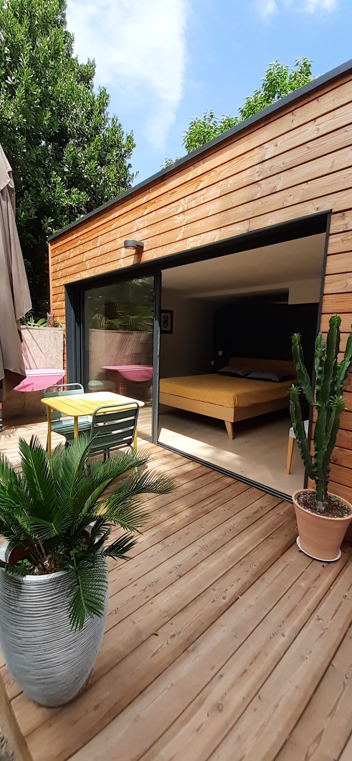 The image showcases a modern wooden room with large glass doors opening to a wooden terrace. A small dining table and chairs are present outside, alongside a potted plant. The spacious bed is visible inside, creating a seamless flow between indoor and outdoor areas.