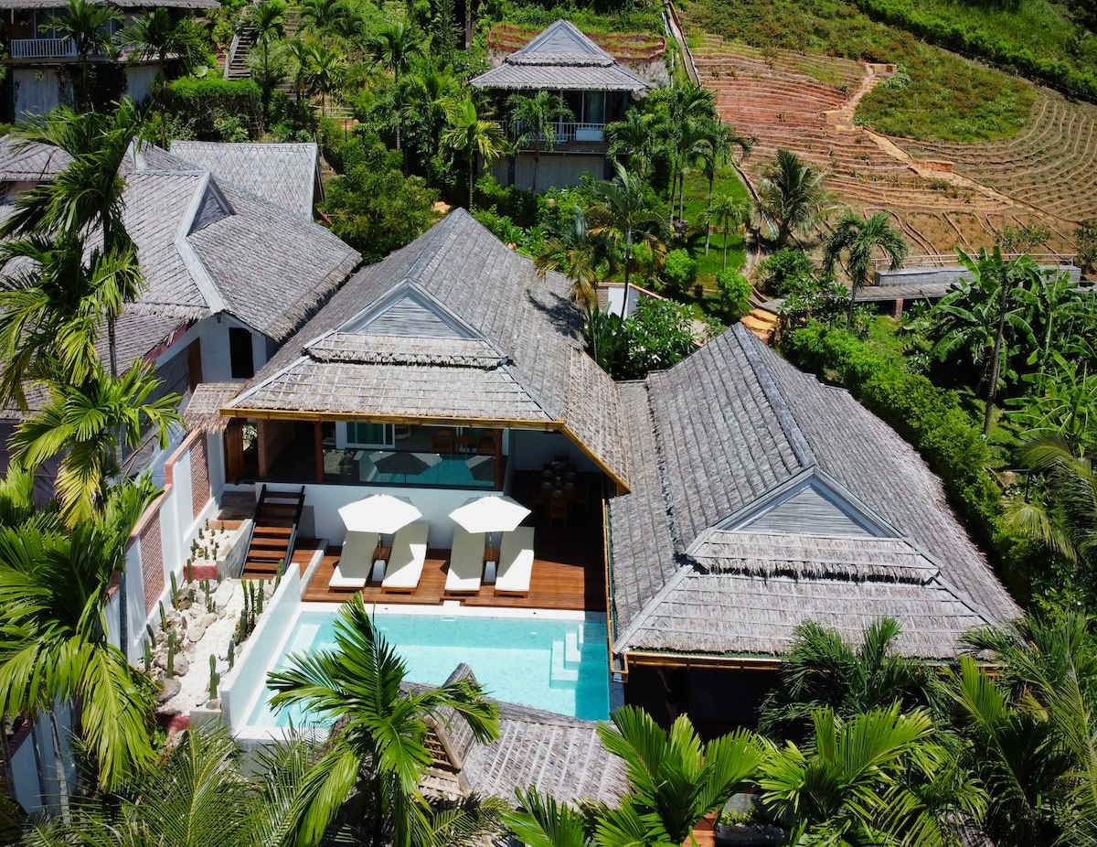 An aerial view displays a tropical villa surrounded by lush greenery. The private pool is bordered by two sun loungers, inviting relaxation. Spacious outdoor decks and thatched roofs characterize the structure, while the backdrop features hints of terraced land and palm trees, enhancing the serene environment.