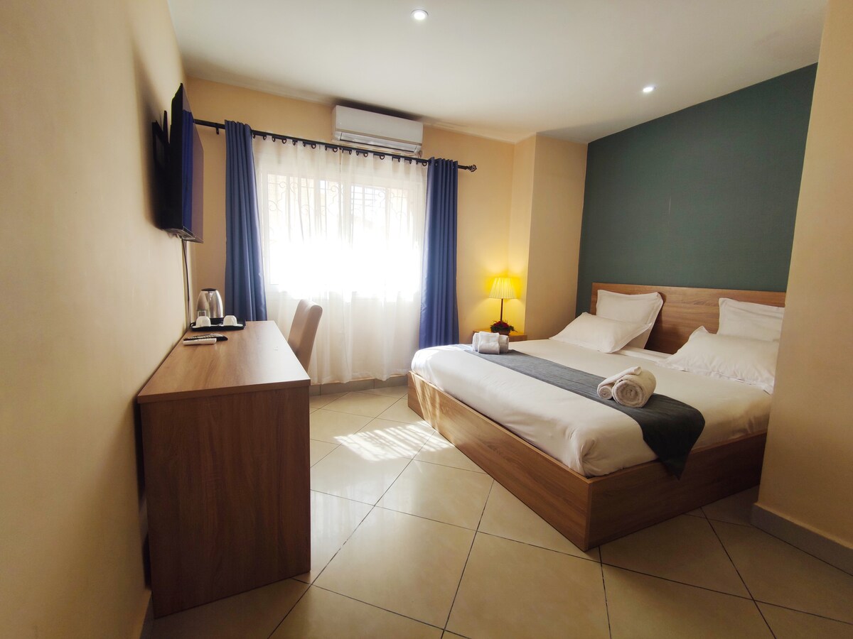 A well-appointed room features a spacious bed with light bedding and a decorative throw. Large windows allow natural light to fill the space, complemented by a small desk and chair. A television is mounted on the wall, providing entertainment options.