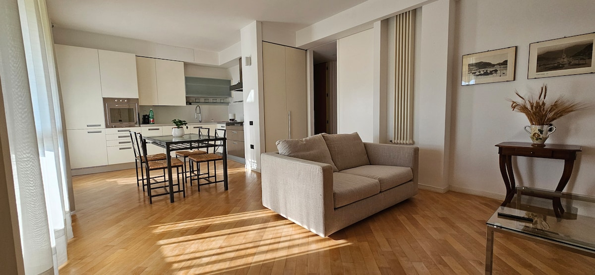An open-plan living area features light wood flooring and modern furnishings. A beige sofa faces a dining table with four black chairs. Light is streamed in through large windows, creating a bright and airy environment. A small side table holds a vase with dried flowers.