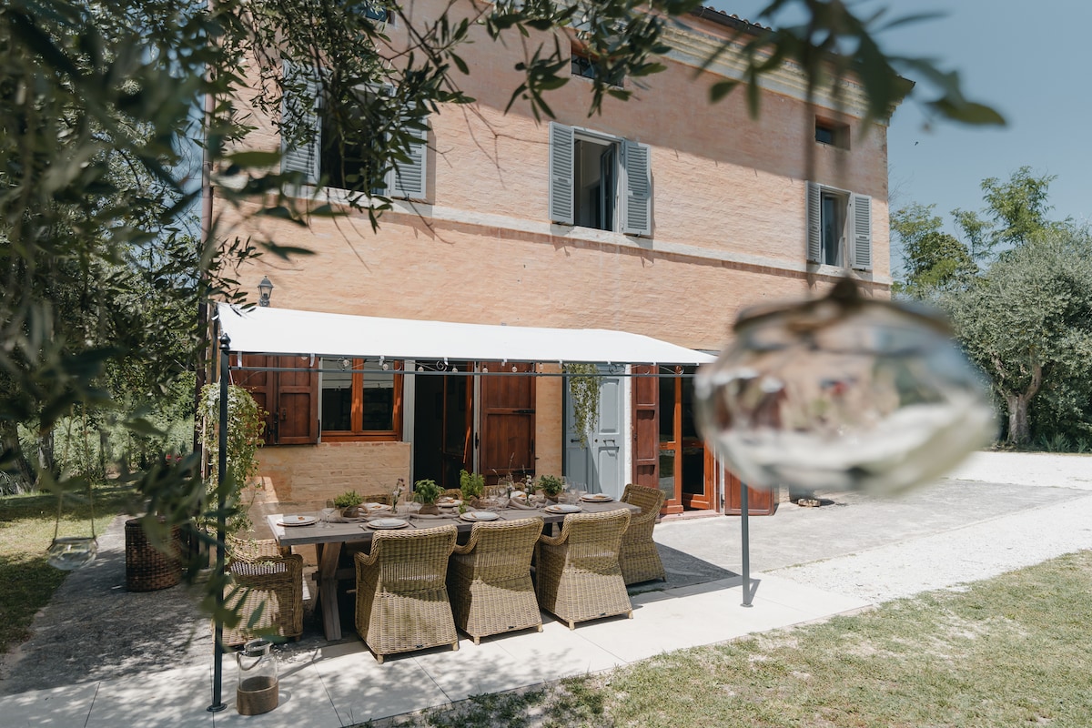 The exterior of the house is shown, featuring a large dining table surrounded by woven chairs under a shaded awning. The warm, brick facade and wooden window shutters create a harmonious blend with the surrounding greenery, enhancing the tranquil outdoor setting.