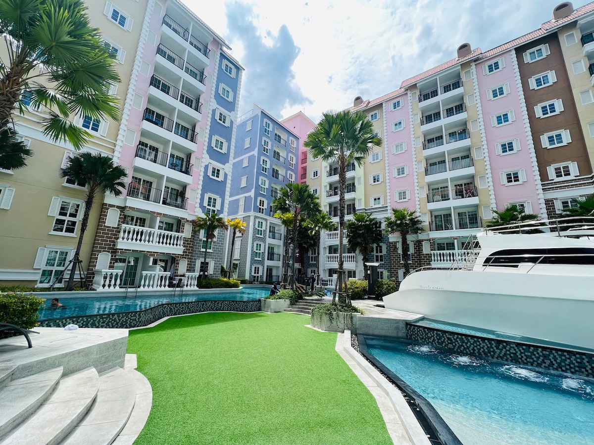 Surrounded by vibrant palm trees, the colorful apartment complex features multiple floors with balconies. A manicured green lawn spans the foreground, leading to a shimmering pool that reflects the sky. A sleek boat is docked nearby, enhancing the relaxing atmosphere.