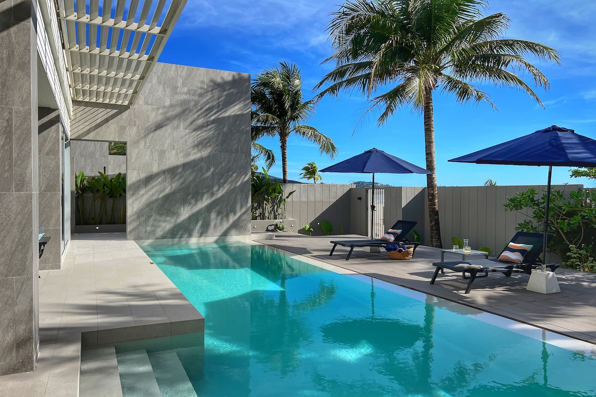A modern outdoor area features a private swimming pool bordered by smooth stone tiles. Sun loungers with umbrellas provide shaded seating options, while tall palm trees add a tropical touch. The clear blue sky enhances the serene atmosphere of the space.