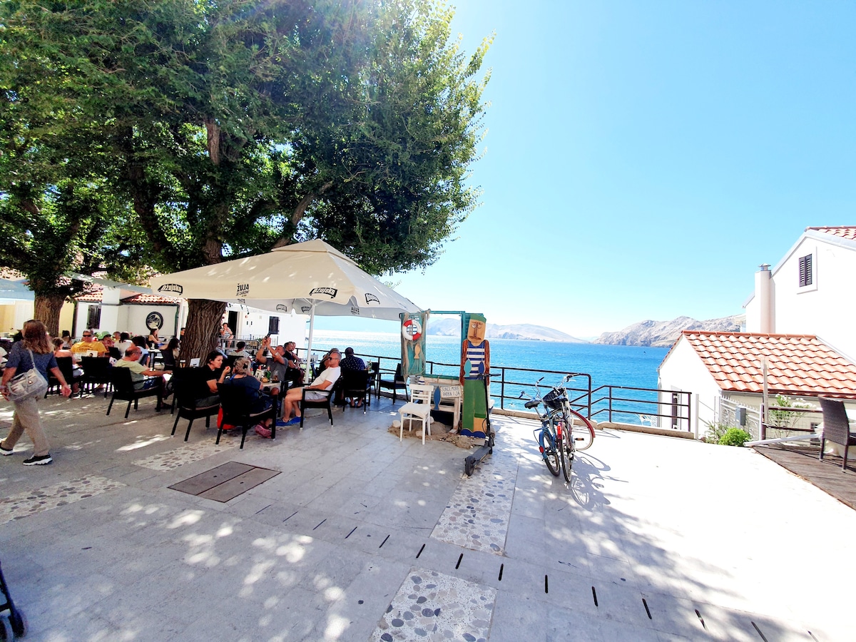 An outdoor seating area is presented along the waterfront, featuring several tables and chairs shaded by a large umbrella. Patrons are seated, enjoying the view of the sea and nearby hills. A bicycle is parked nearby on the stone pathway.
