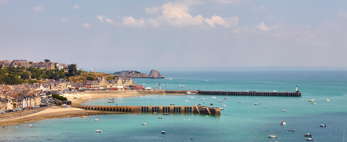 The image captures a panoramic view of Cancale's coastline, featuring a calm bay dotted with boats. Sandy shores are lined with houses, while a harbor extends into the water. The scene is accented by gentle waves and a clear blue sky, enhancing the coastal ambiance.