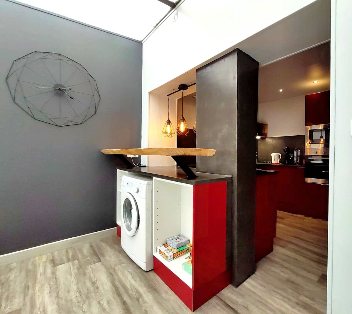A modern kitchen area features a sleek design with red cabinetry and a contrasting dark countertop. A washing machine is integrated into the cabinetry, and pendant lights provide ambient lighting. The open layout connects to the living space, enhancing the overall functionality.