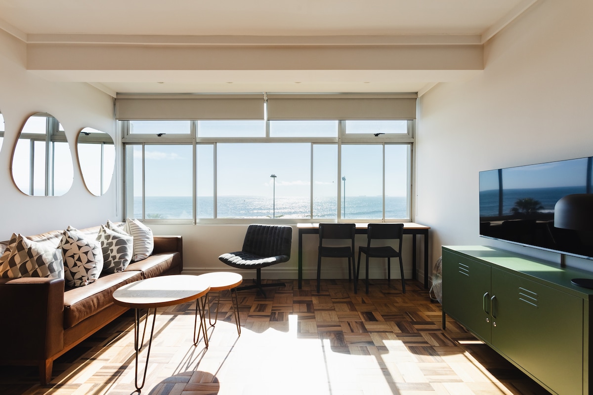 A spacious living area features large windows offering views of the ocean. Comfortable seating includes a leather sofa and a sleek chair. A round table rests on a wooden floor, complemented by a minimalist television unit. Natural light fills the room, creating an inviting ambiance.