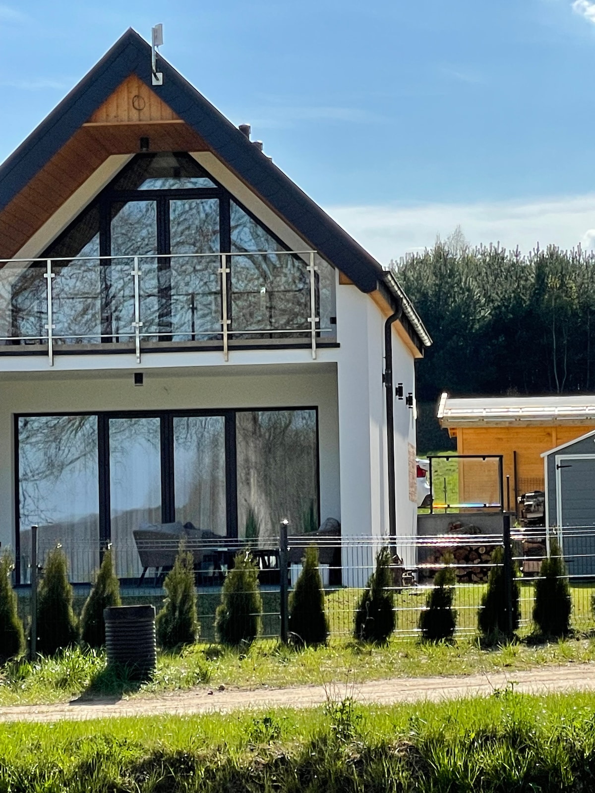 A modern house features large glass windows and a balcony, presenting a contemporary architectural style. The exterior is complemented by a sloped roof, while a neatly arranged row of small manicured shrubs lines the foreground. A wooden structure is visible in the background.