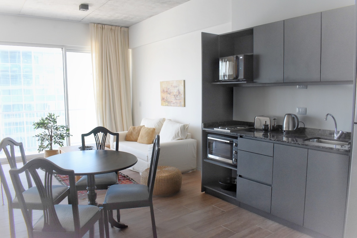 A modern kitchen area is integrated with a dining space featuring a round table and four chairs. A comfortable seating arrangement is visible in the background, along with large windows allowing natural light to fill the room.