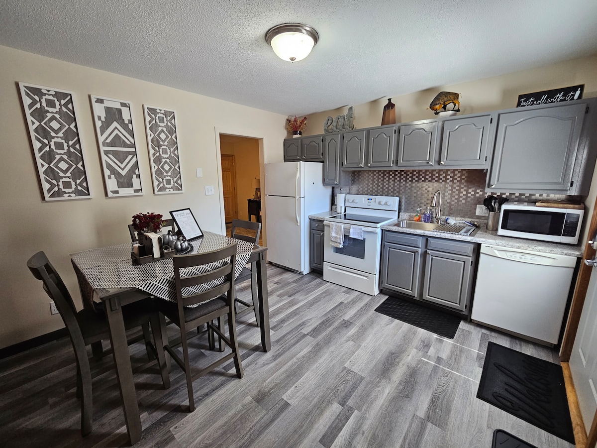 A fully equipped kitchen is shown, featuring grey cabinetry and modern appliances. A dining table with four chairs sits adjacent to the kitchen area. Wall art is displayed above the table, enhancing the inviting atmosphere. Natural light enters through a nearby doorway.