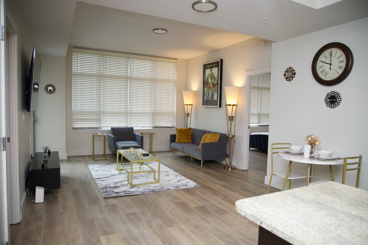 A modern living area is highlighted by large windows allowing natural light to fill the space. A comfortable gray sofa and stylish armchairs are arranged around a decorative area rug. A dining table for two is positioned against the wall, complemented by elegant wall decor and a clock.