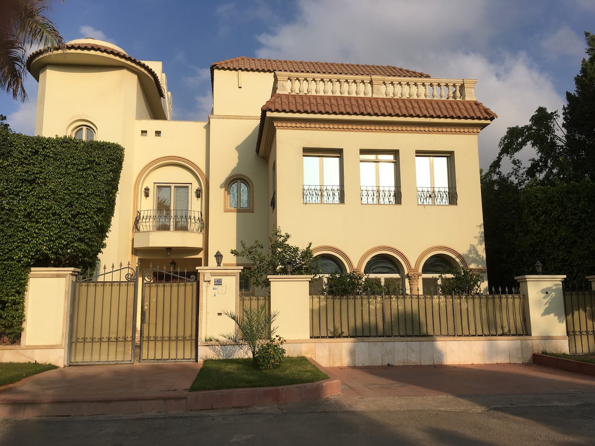 The exterior of a spacious two-story home is showcased, featuring a light yellow facade with ornate architectural details. Large windows allow for generous natural light, while manicured greenery lines the gated entrance, providing a sense of privacy and security.