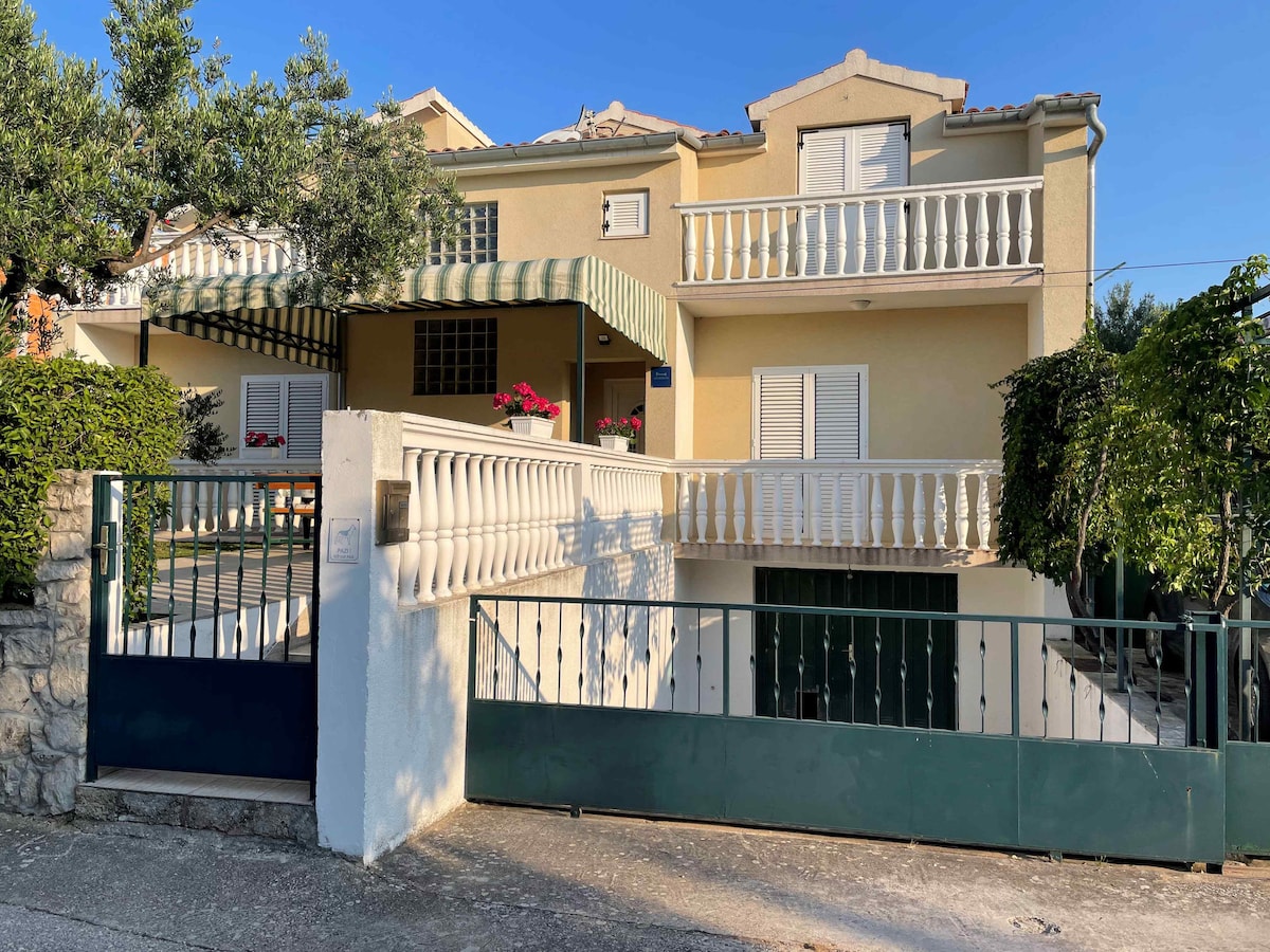 An inviting entrance to a two-story family house is framed by lush greenery. The structure features a covered balcony with striped awnings and flower boxes. A gated pathway leads to the entrance, ensuring a peaceful and private atmosphere.