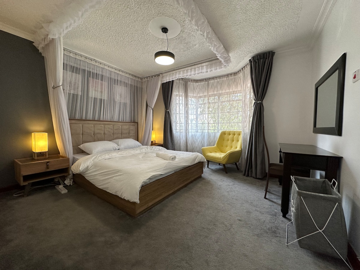 A spacious bedroom features a large bed draped with a canopy. Soft lighting is provided by bedside lamps and a ceiling fixture. A cozy armchair sits by the window, which is adorned with sheer and dark curtains. A mirror and a small desk complete the room's design.