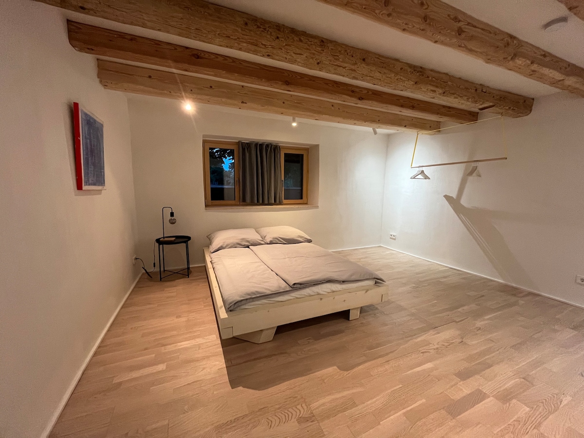 A sleeping area features exposed wooden beams and a light wooden floor. A spacious bed is accompanied by a small nightstand, with a reading light positioned nearby. Natural light enters through the window, which is adorned with light curtains that can be drawn for privacy.