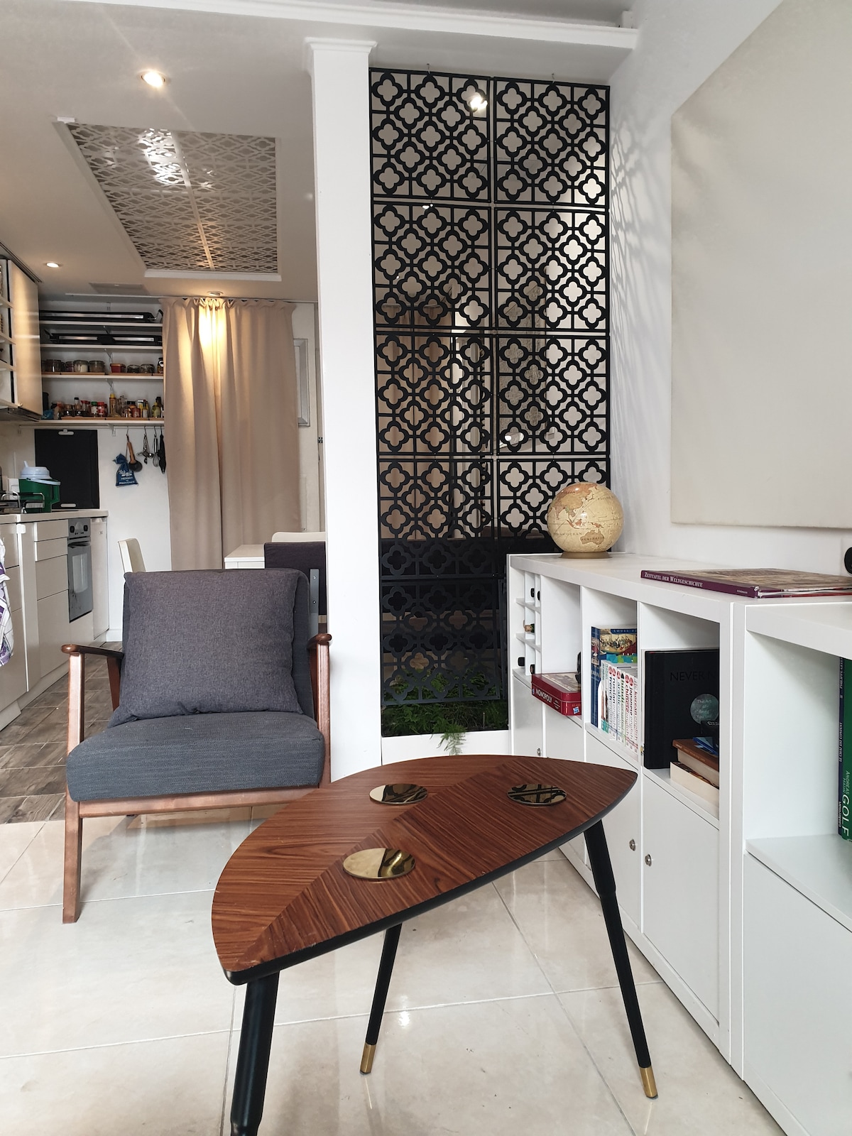 A cozy corner is depicted, featuring a stylish wooden accent table with decorative objects. A comfortable grey armchair is seen, complemented by a partial view of a modern kitchen in the background. Decorative screens create an intimate ambiance, enhancing the space's modern design.