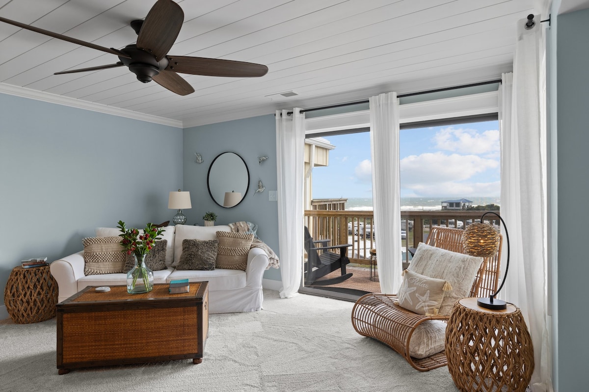 A comfortable living area is present, featuring a light-colored sofa and a rattan chair. Natural light filters through sheer curtains, illuminating the space. A coffee table, accompanied by decorative elements, anchors the room. Sliding glass doors lead to a balcony with ocean views.