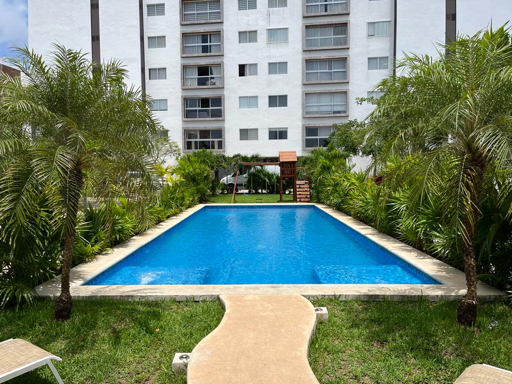 A clear blue swimming pool is framed by lush green landscaping, including palm trees and ornamental plants. A pathway leads to the pool area, providing easy access for guests. The backdrop features a multi-story building with numerous windows.