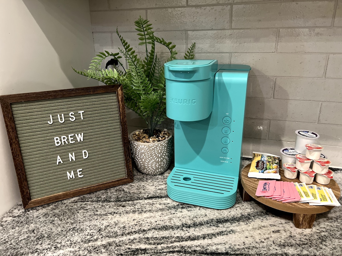 A light blue Keurig coffee maker is positioned on a granite countertop alongside a variety of coffee pods and creamers. A decorative sign with the phrase 'Just Brew and Me' is displayed next to a potted plant, adding a personal touch to the space.