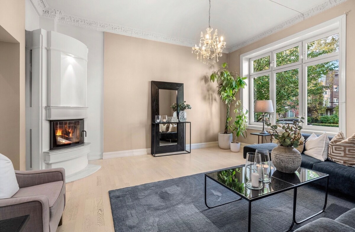 A spacious living area is characterized by bright natural light flooding through large windows. A modern coffee table is centered on a soft rug, surrounded by comfortable seating. A decorative mirror and a plant add subtle accents, while a stylish chandelier hangs from the ceiling.