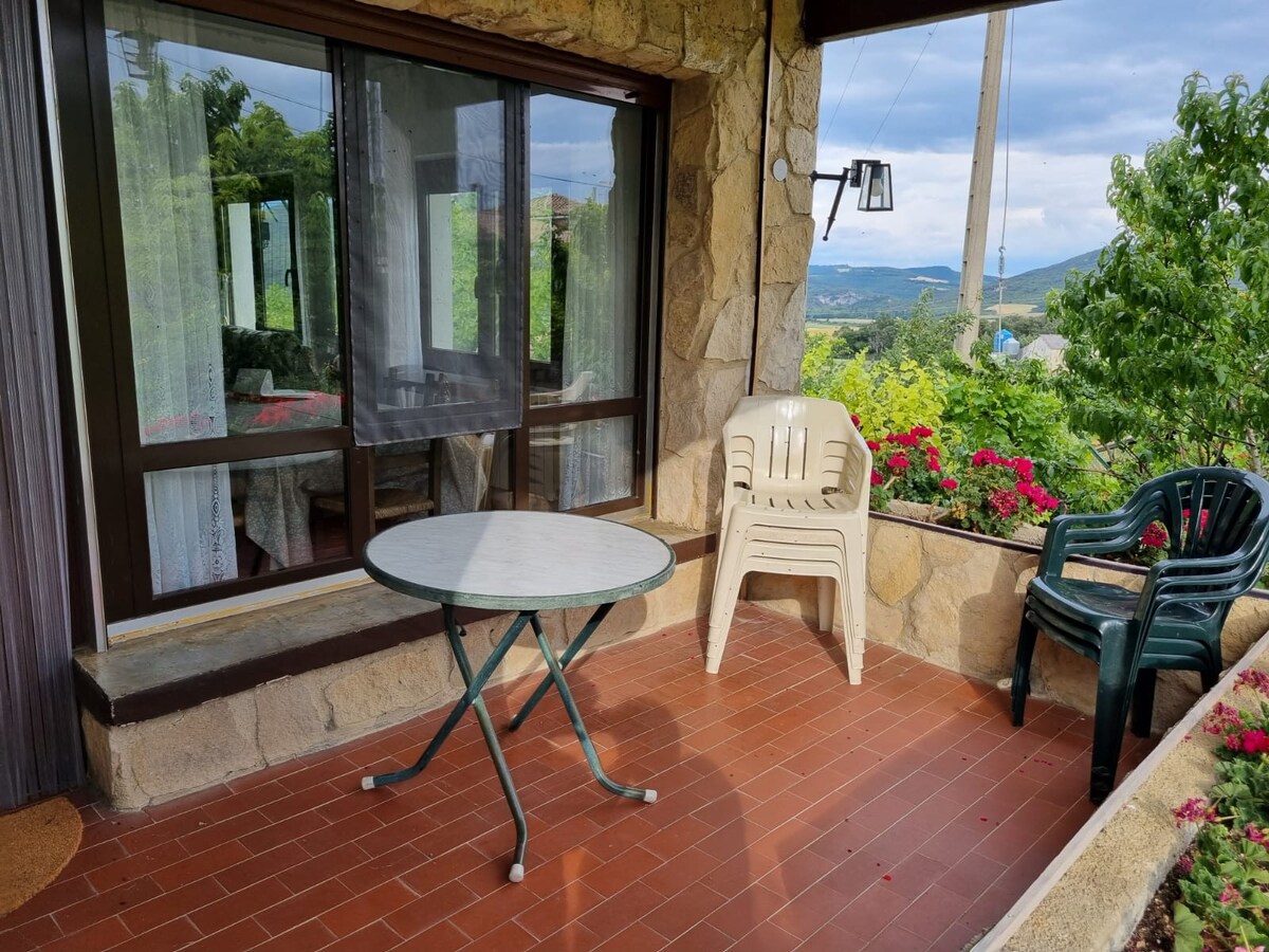 A shaded terrace features a round table accompanied by a green chair and a stacked plastic chair. Flower boxes adorn the edges, adding color to the stone structure. Large windows provide views of the natural surroundings, creating an inviting outdoor space.