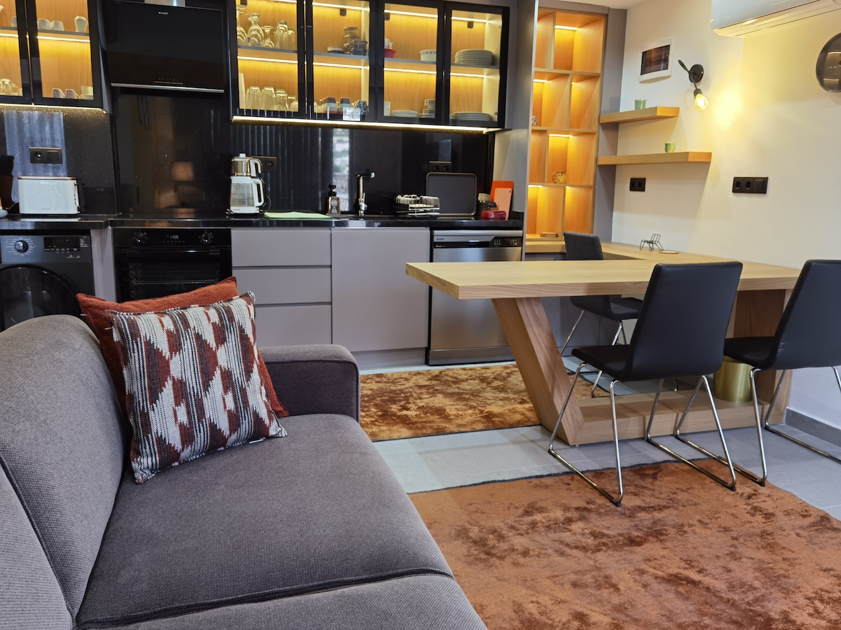 A modern kitchen and dining area is displayed, featuring sleek cabinetry and a warm ambient glow. A wooden dining table, accompanied by black chairs, contrasts with the soft tones of the rug. A comfortable gray couch with a patterned pillow enhances the relaxed atmosphere.