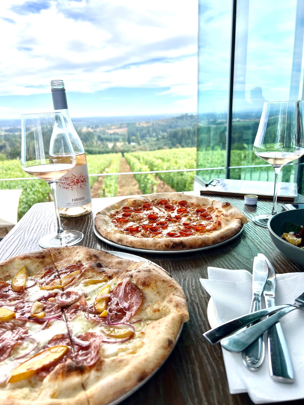 Two pizzas are displayed on a wooden table, one topped with cured meats and yellow fruit, the other with ripe tomatoes. Two wine glasses and a bottle of rosé are nearby, with a scenic vineyard visible through large glass panels, reflecting a clear sky.