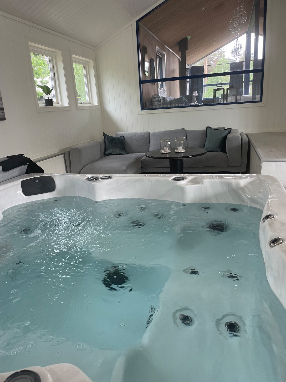A soothing spa area is showcased, featuring a hot tub filled with clear water. A spacious seating area with a sectional sofa and two side tables is visible in the background, adorned with decorative cushions. Light enters through large windows, enhancing the tranquil environment.