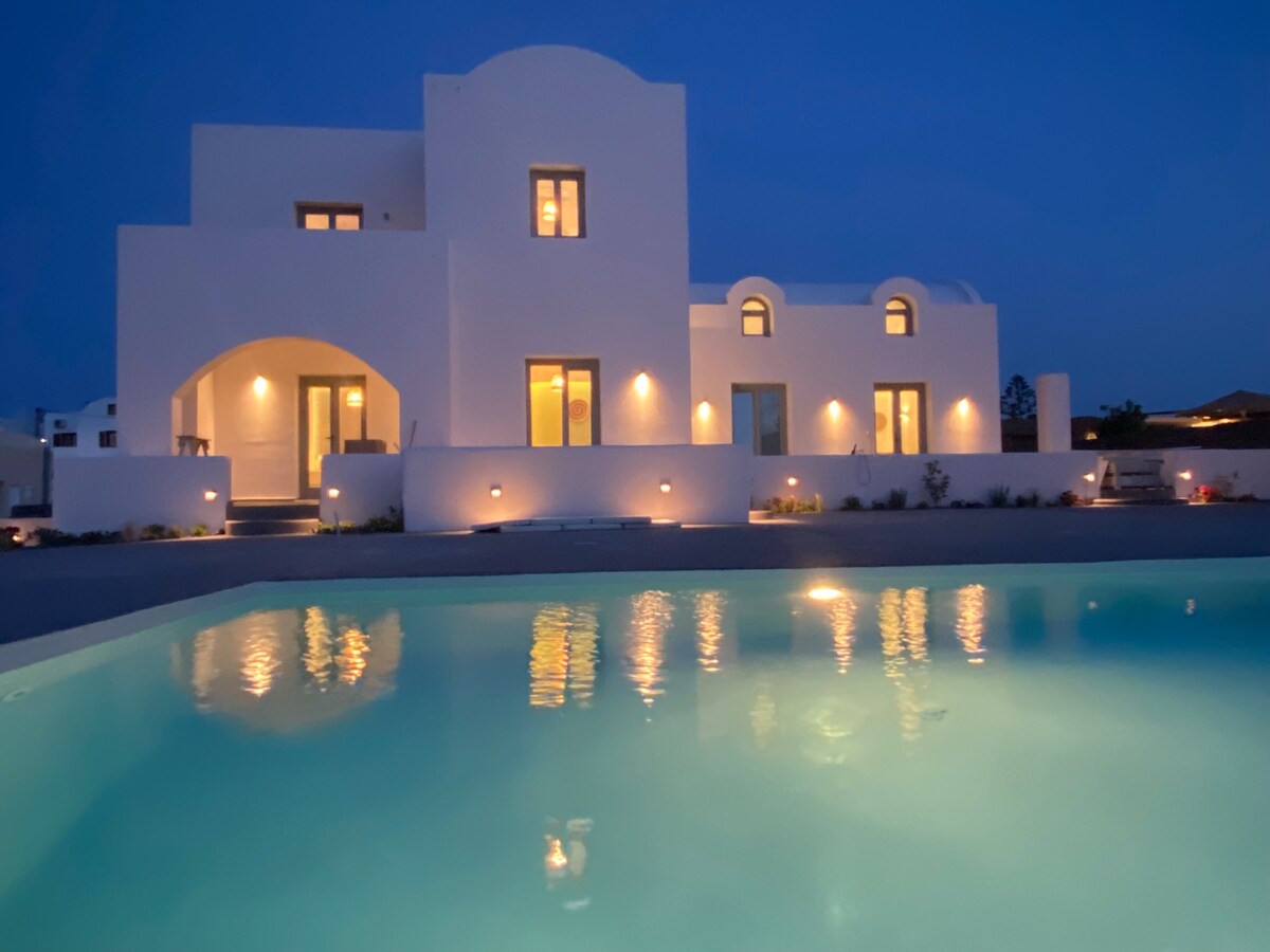 The exterior of the property is illuminated at night, reflecting softly in the clear water of the pool. The white structure features multiple windows, highlighting the contemporary architecture. Surrounding landscaping includes minimalistic plantings, enhancing the serene ambiance.
