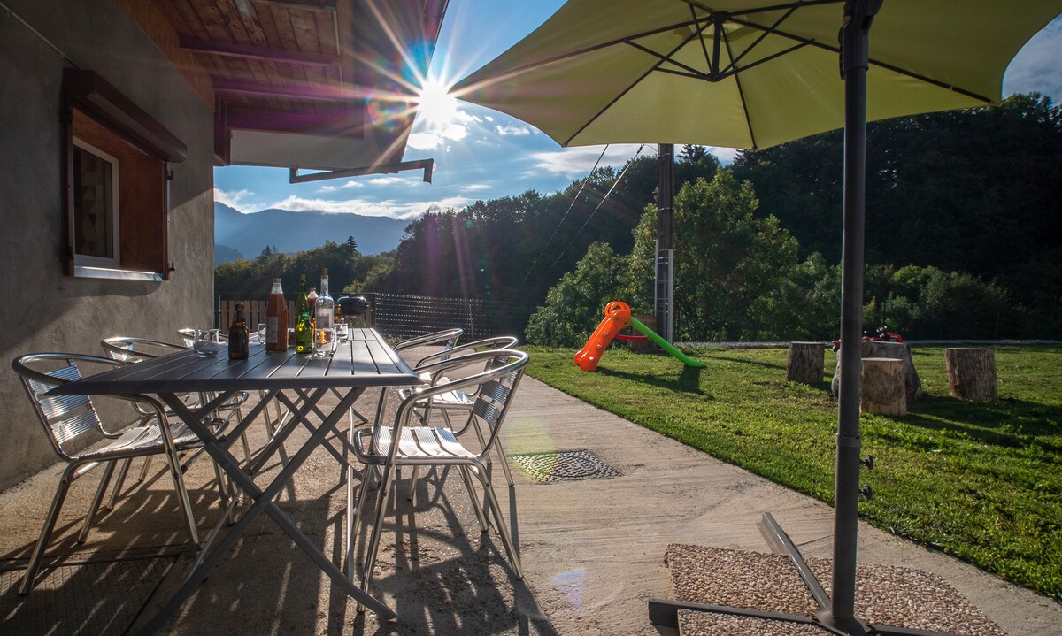 An outdoor terrace is presented with a large table and metal chairs, surrounded by green grass. A sunshade offers protection from the sun. In the background, a slide stands among the trees, while distant mountains add to the serene atmosphere.